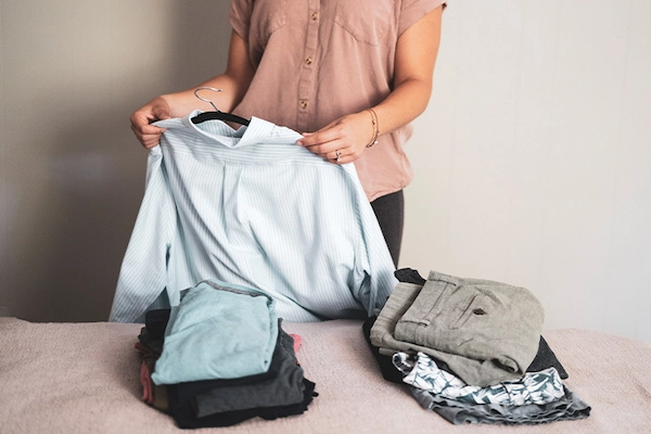 Person holding up and inspecting a blue button down shirt with folded clothes on bed in foreground.